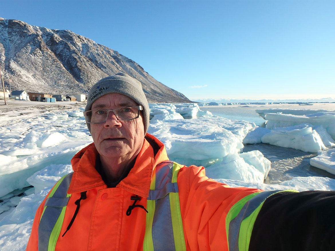 Autoportrait de Klaus Dohring, qui porte un manteau de chantier orange, avec la communauté de Grise Fiord en arrière-plan.