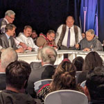 Dignitaries sitting onstage at a table and the audience watch as NTI President Aluki Kotierk signs the Nunavut Lands and Resources Devolution Agreement.