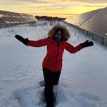 A participant standing in the snow with arms outstretched, gesturing toward long arrays of solar panels on either side.