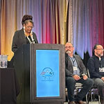 Tracey Willoughby standing behind a podium onstage. Other speakers sit in a row to the right.