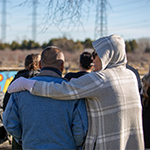 Two people standing at the back of a group, with their backs to the camera. One person has their arm around the other’s shoulder.