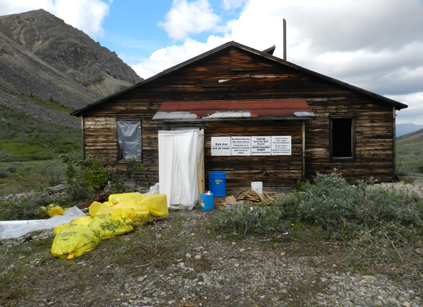 An old wooden building along the Canol Trail. White tarps mark off an area that's been decontaminated. Yellow hazardous waste bags are stacked in front of the building. Mountains are in the background.