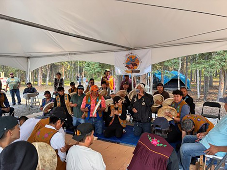 Cette photo montre des membres de la Première Nation des Dénés de Yellowknife agenouillés en deux groupes, face à face, sous l'auvent d'une tente. Beaucoup d'entre eux jouent du tambour.