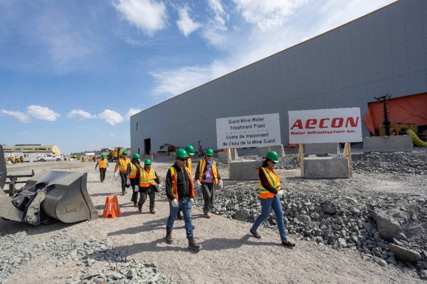 A group of people walk along a gravel path beside an excavator outside a large industrial building.