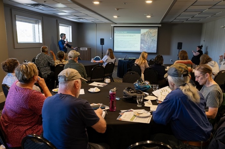 Several people seated at round tables listening to a presenter at the front of the room, a PowerPoint slide visible on a screen.
