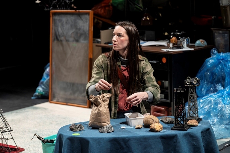 A woman with long dark hair and a flannel shirt sits at a table examining rocks as part of a play.