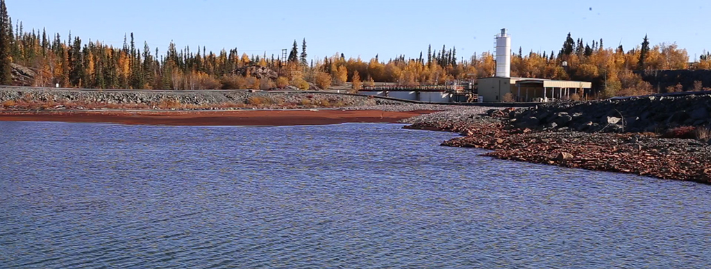 This picture shows the Giant Mine effluent treatment plant from across a body of water. The plant is a group of buildings and large basins for water treatment. Trees are behind the building and along the shore. Rust-coloured water, from the arsenic treatment, is seen on the shoreline, separate from the clean water in the foreground.