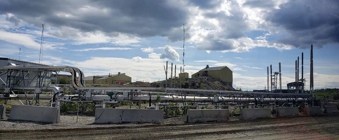 A developed industrial area of the Giant Mine site showing vertical pipes and thermosyphons, which are long vertical pipes filled with carbon dioxide. Buildings are visible on a rocky outcrop in the background of the photo.