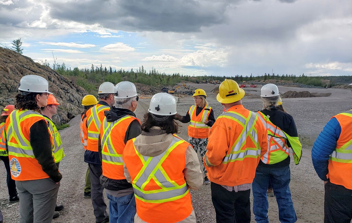 Un groupe de personnes portant des gilets de sécurité et des casques de sécurité se trouve debout sur une route de gravier avec un affleurement rocheux et des arbres visibles en arrière-plan.