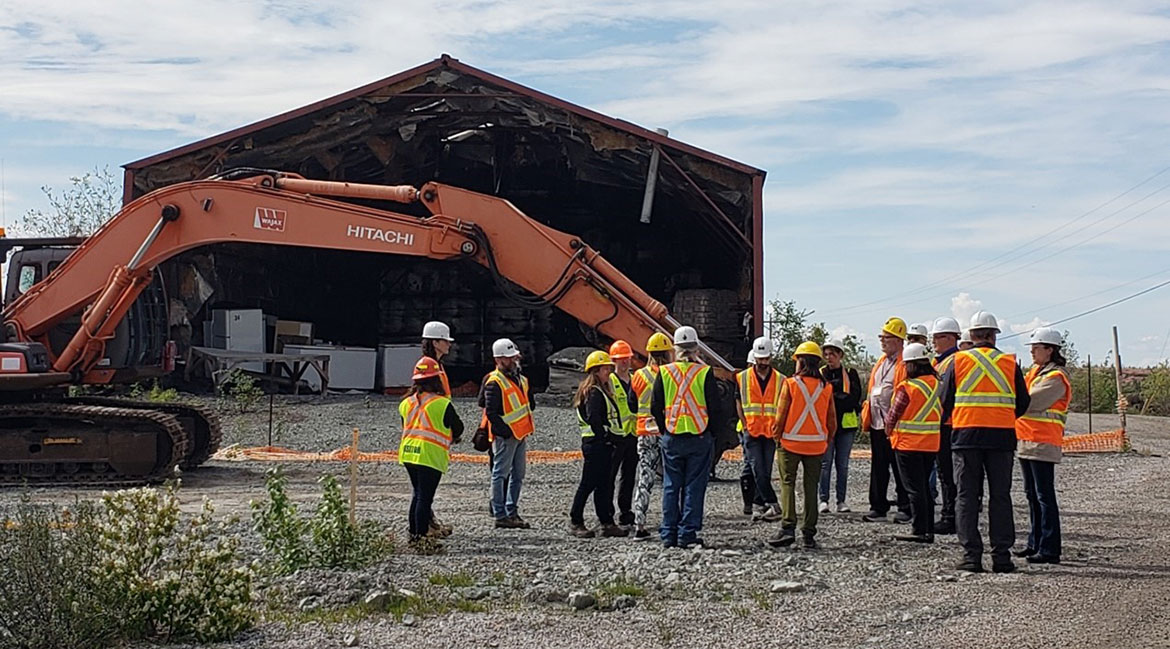 A groups of people in safety vests and hard hats stands next to an excavator. In the background, a building frame with stored debris beneath is visible.