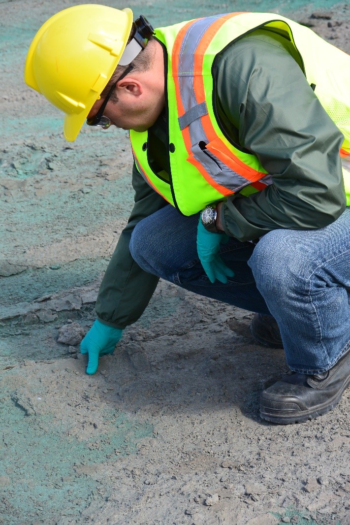 A person wearing personal protective equipment that includes a hard hat, safety glasses, a reflective vest, gloves, and safety boots examines a layer of hardened dust on top of tailings.