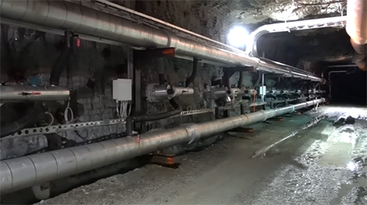 Vue d'une chambre de mine souterraine montrant un sol humide et des tuyaux courant le long du côté gauche.