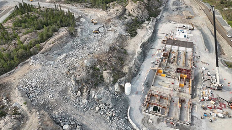 An ariel view of a construction site showing a building foundation on the right and blasted rock with an excavator and a dump truck on the left.