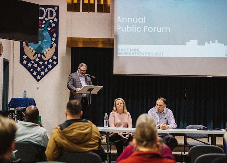 A man speaks at a lectern alongside a panel seated to his right. Above them is a projection screen showing a presentation.
