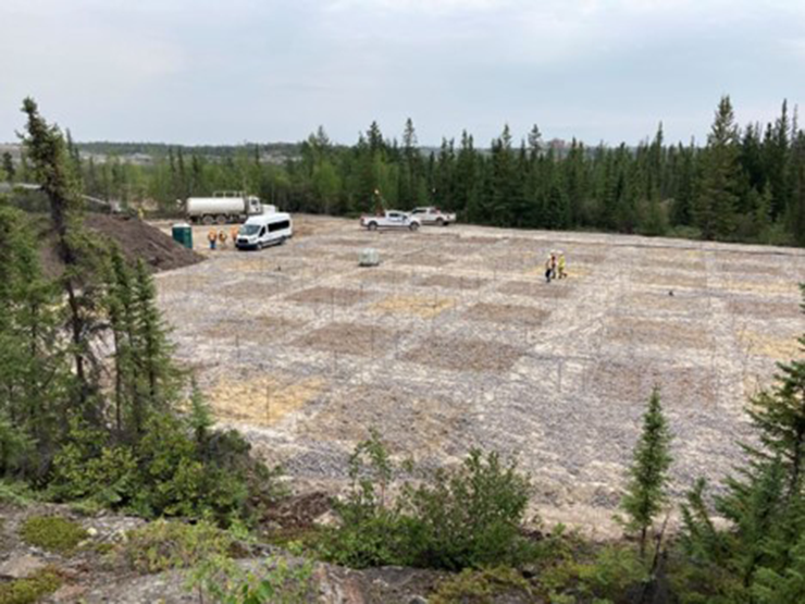 Three construction site workers in personal protective equipment walk across a soil grid surrounded by trees with work vehicles and other workers in personal protective equipment visible in the background.
