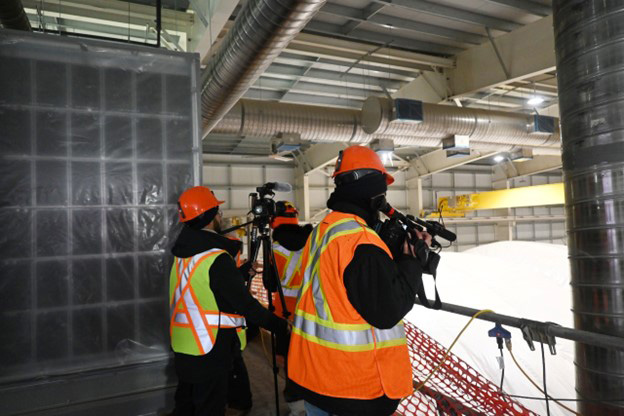 Three people in hard hats and safety vests take photo and video footage from inside a building that is still under construction.