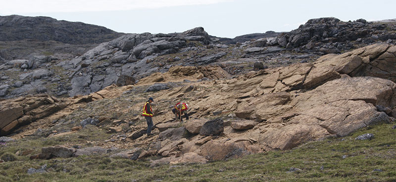 2 geologists on a large, beige-coloured outcrop of bedrock on the side of a hill. 1 geologist is collecting a rock sample