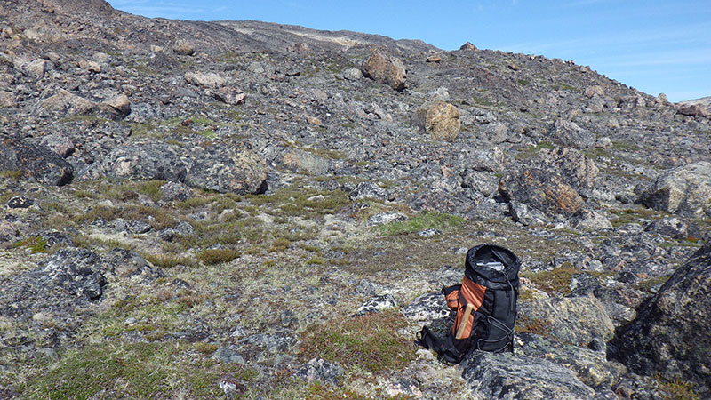 A geologist's backpack, with a sledgehammer handle visible in the side pocket, sitting on a rocky tundra hillside on a sunny day