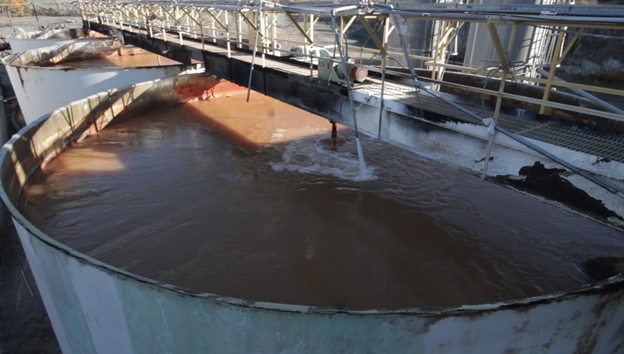 This picture shows a large vat of water being treated at the current seasonally-operated Effluent Treatment Plant at the Giant Mine site. The water is a red-brown colour from the ferric sulfate used so arsenic and other metals will settle out of the water. A metal catwalk is visible over the large vat.