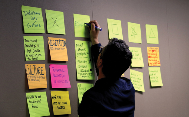 A consultant is shown writing on one of the many pieces of paper stuck to a wall, representing categorized concerns about the Giant Mine site.