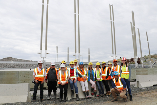 A group of fourteen people dressed in personal protective equipment on the Giant Mine site stand in front of the thermosyphons, which are long vertical pipes sticking out of the ground.