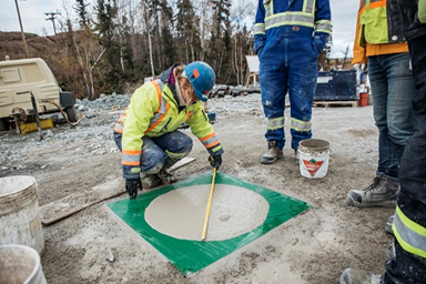 Image shows a woman in personal protective equipment measuring a concrete sample at Giant Mine, while three workers in personal protective equipment observing the testing can partially be seen.