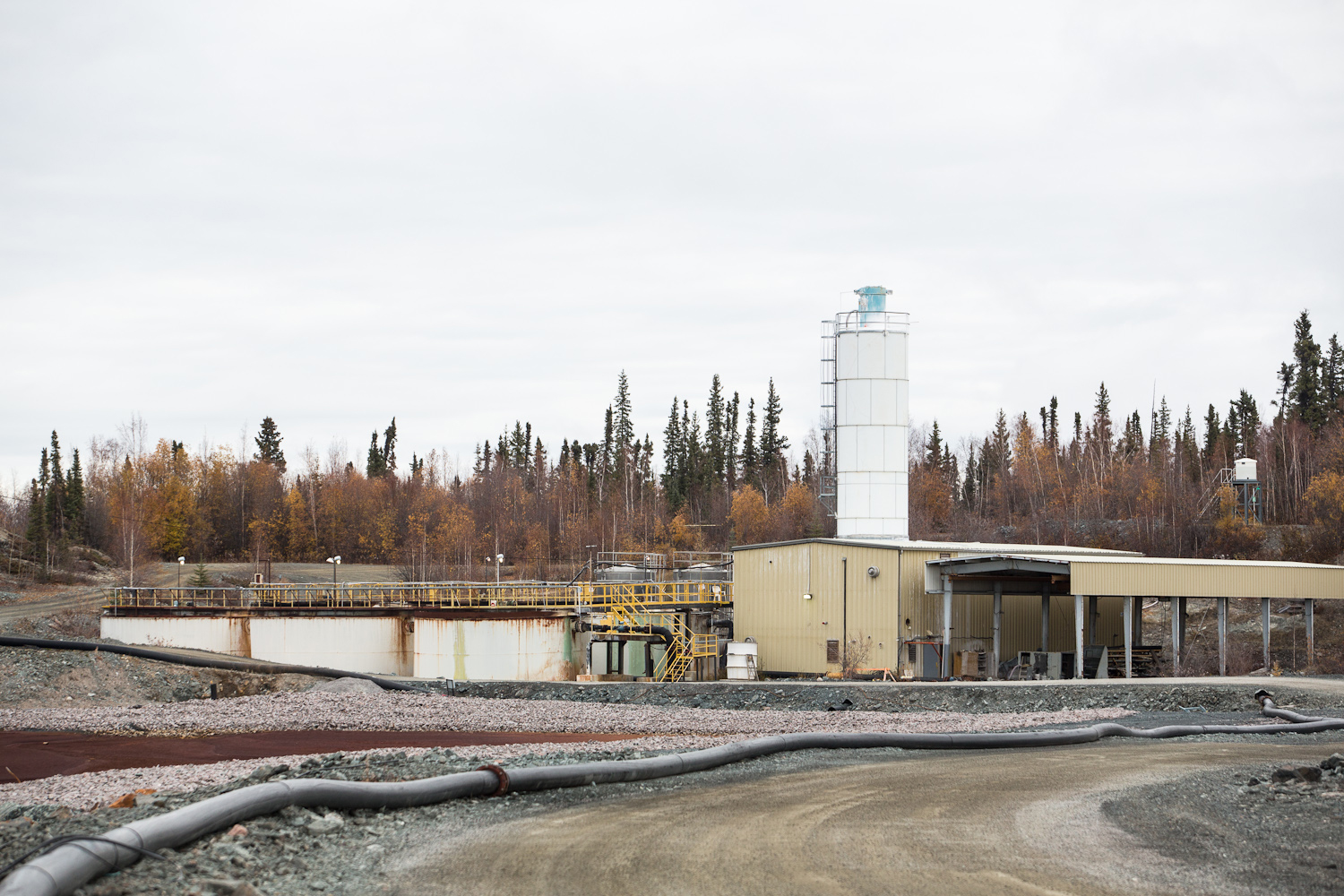 La photo montre un tuyau menant à une usine de traitement des effluents. Trois grands réservoirs sont visibles à côté d'un bâtiment, avec des arbres en arrière-plan.