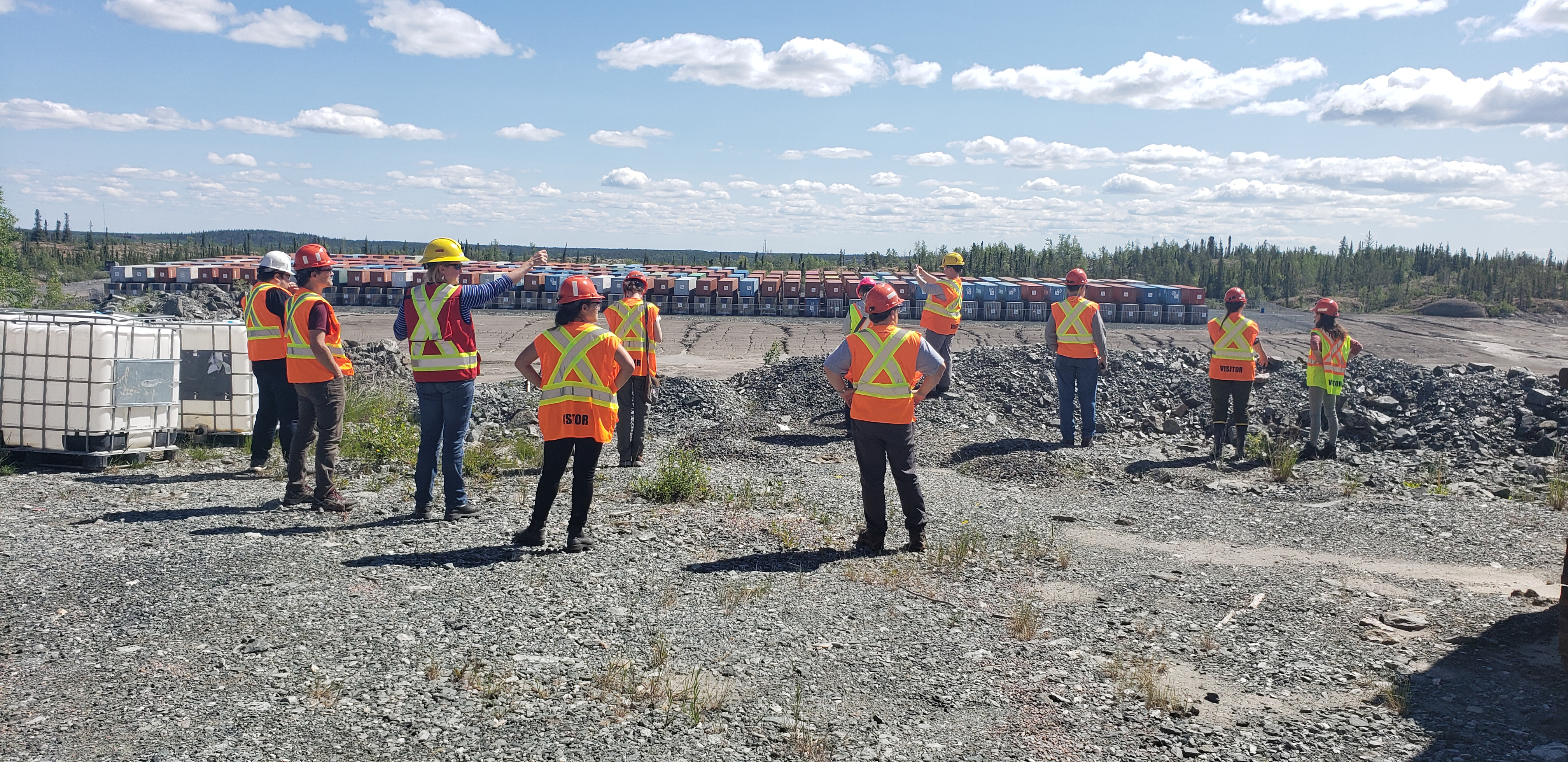 Eleven people in personal protective gear look over a grey, rocky area on which hundreds of sea cans are stacked.