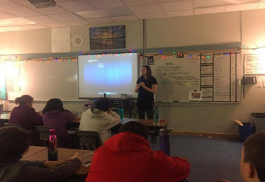 This picture shows a woman next to a presentation screen in front of a class of junior high students.