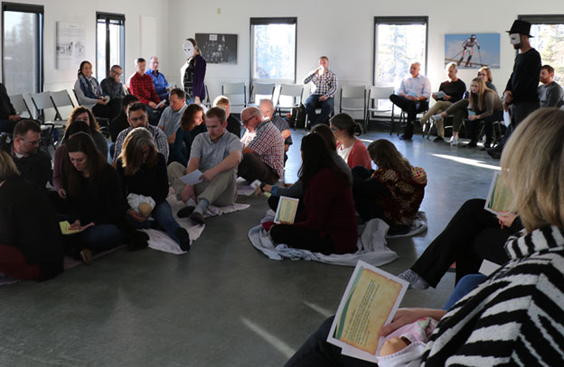 This picture shows Project team members participating in the Blanket Exercise. Some members, representing Indigenous peoples, sit on the floor on blankets that represent Indigenous lands, which were made smaller and divided throughout the exercise. Other members sit in chairs in a circle around the blankets, as they represent Indigenous people who have died and have been removed from the exercise. Two people in white masks, who represent colonizers, circle around the seated team members on the blanket.