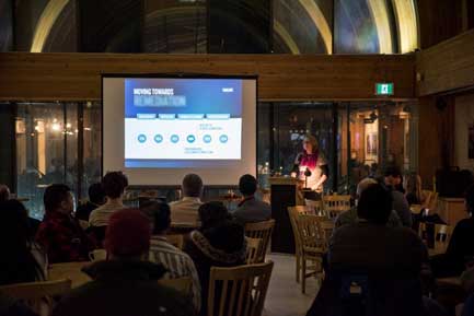 This picture shows people sitting at tables facing a presentation screen. The words "Moving Towards Remediation" are visible on the screen. A woman stands at a podium, facing the crowd as she presents.