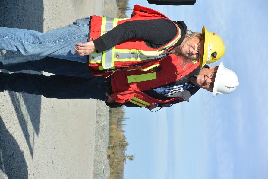 A woman and a man, both smiling, in personal protective equipment on a flat gravel surface on the Giant Mine site.